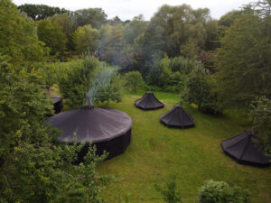 Zwei Jurten und drei Kohten stehen auf einer Lichtung zwischen Bäumen.